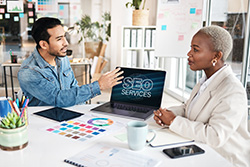 Ocean County Web Design professional meeting with client over design materials and a laptop open wiith a screen displaying "SEO SERVICES"